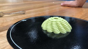 A puree food shape is resting on a black circular mat. The shape is pale green as it is made of guacamole. The shape is layered and tapers up, wider at the base than at the top. 
